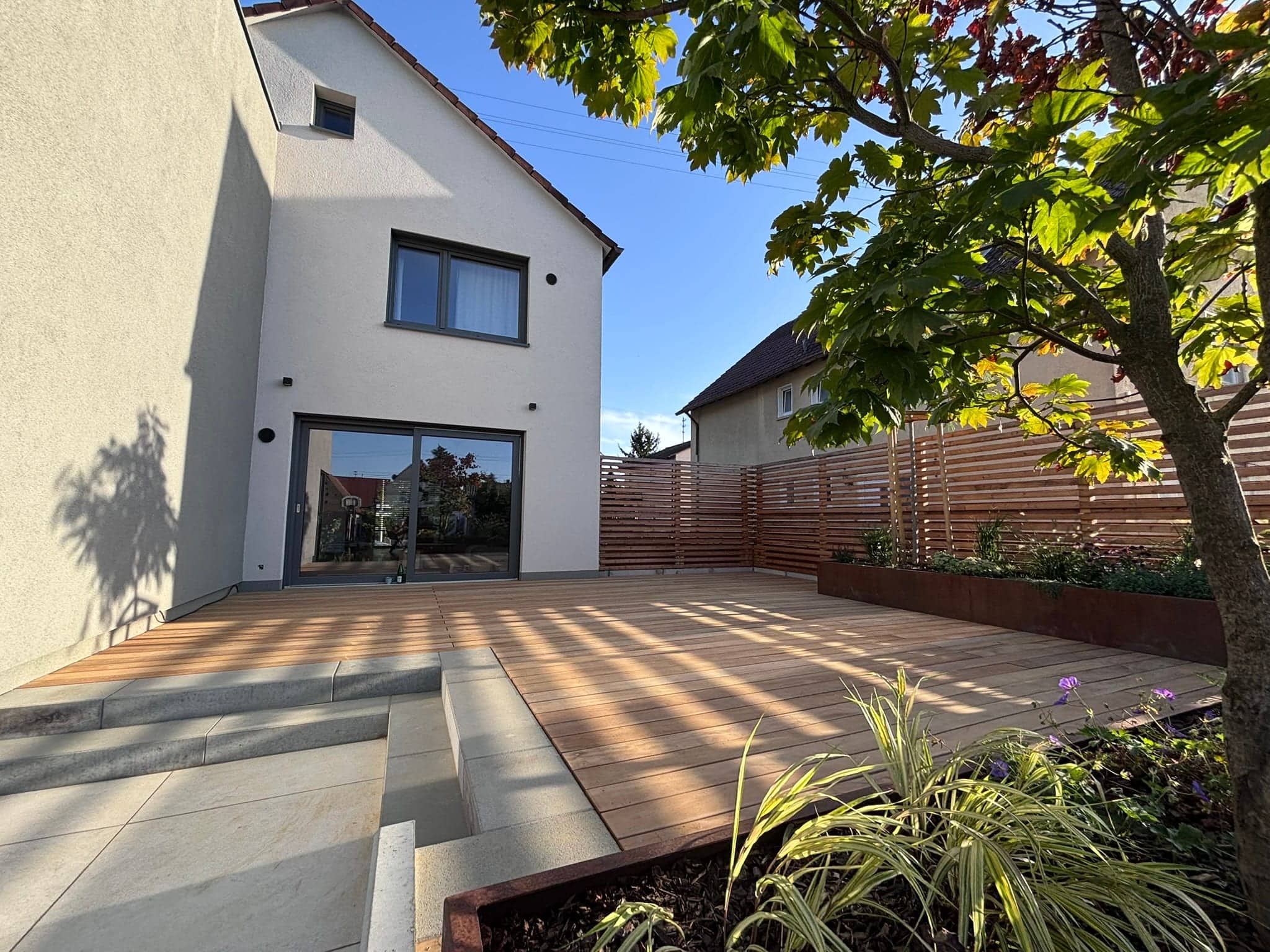 Holzterrasse mit Betonstufen und Pflanzen vor einem hellen Einfamilienhaus
